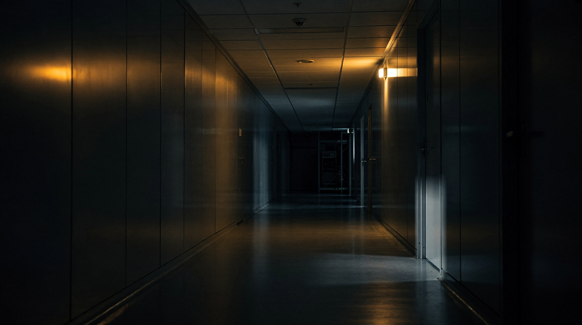A dark laboratory corridor, one fluorescent tube lit near the ceiling, the far door barely visible.