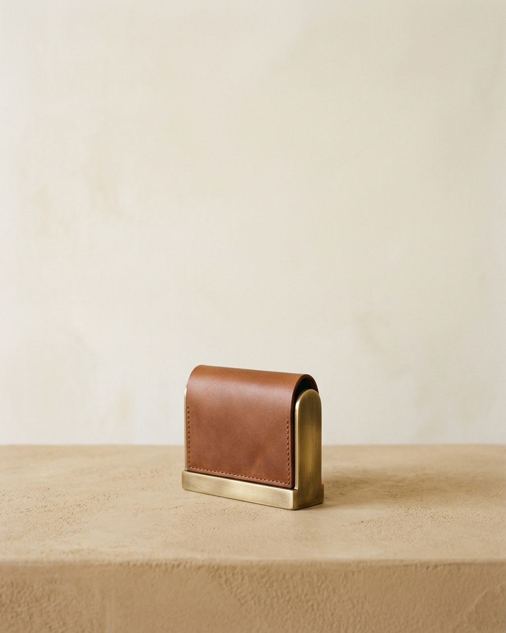A cognac leather card case standing in a brushed brass cradle on a sand-coloured stone bench against a plaster wall