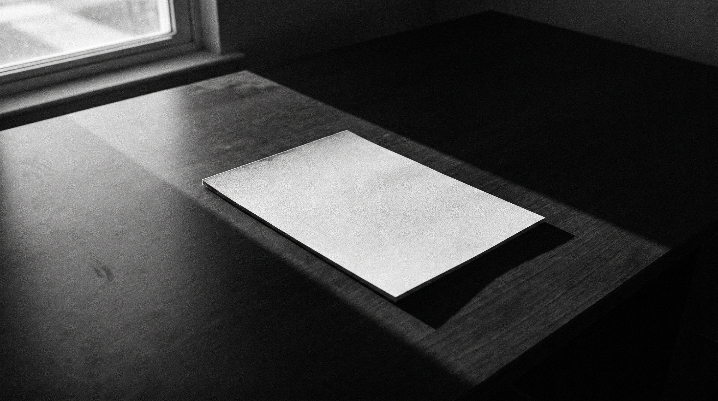 A single blank sheet of heavy paper on a dark desk, one shaft of window light falling across it.